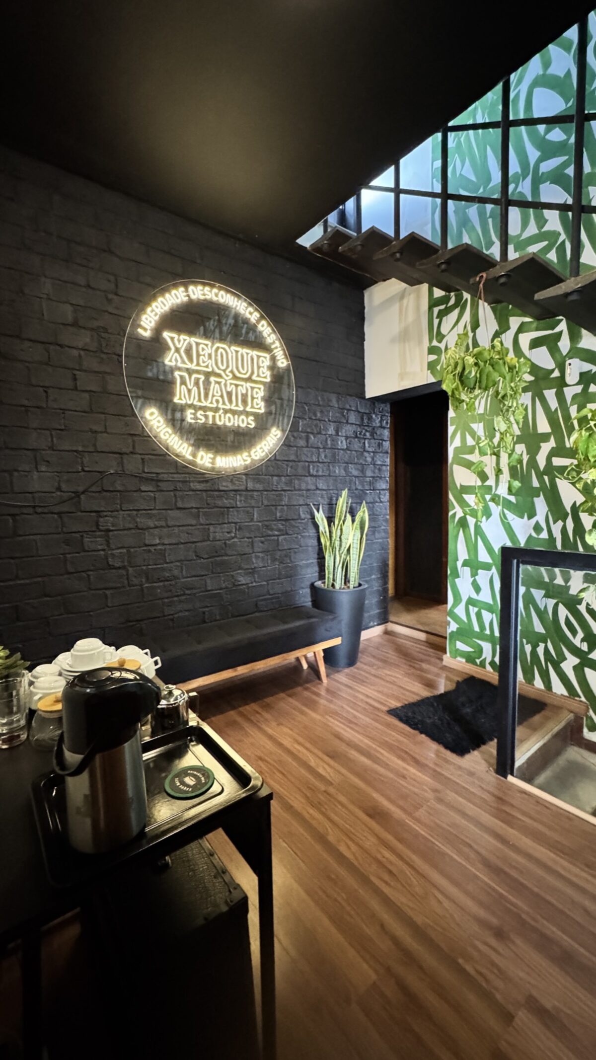 Entrada do estúdio: parede de tijolo preto com o neon Xeque Mate Estúdios e a parede de caligrafia verde